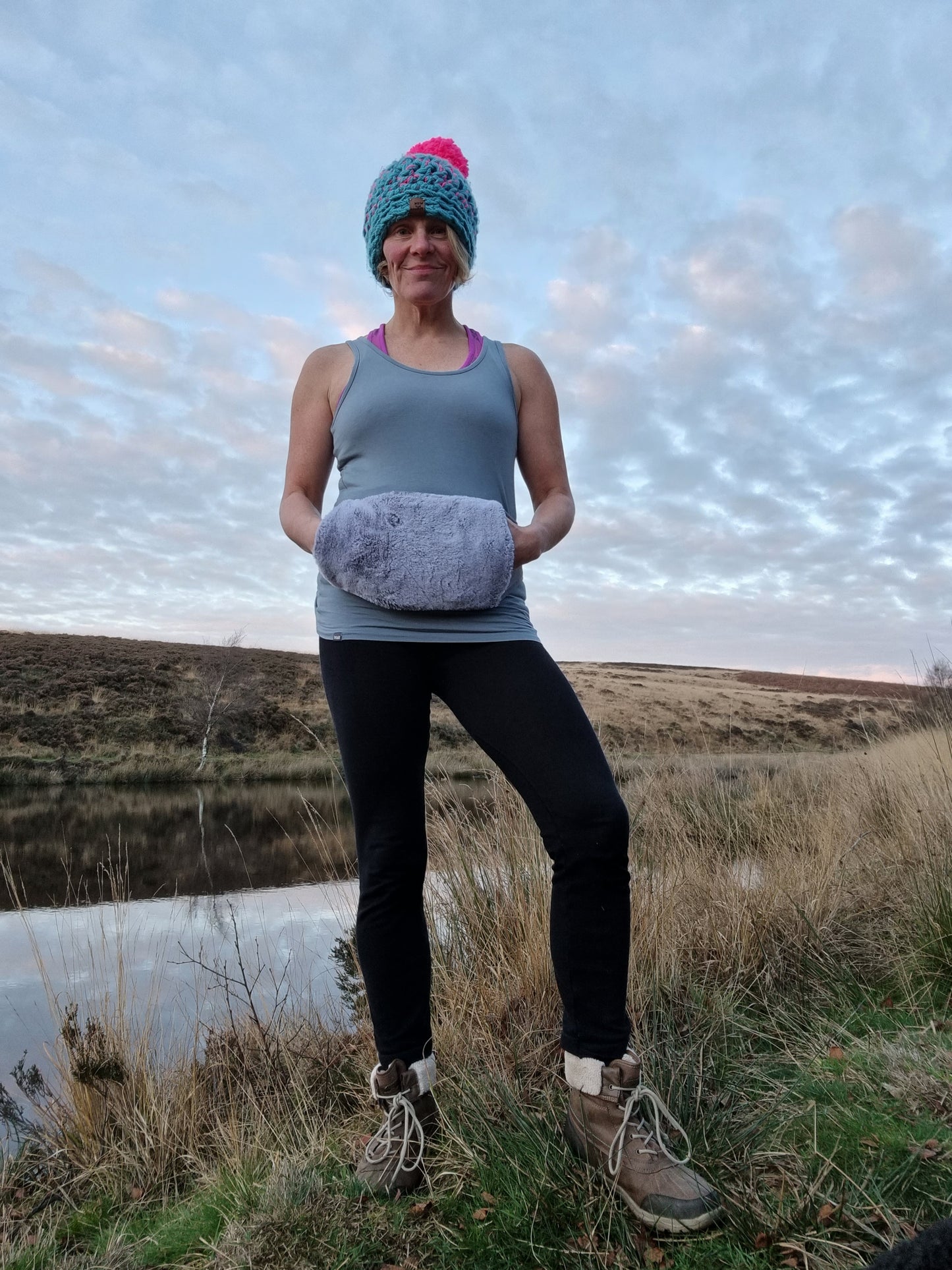 a lady stands in front of a lake and has her hands warming in a Hottum core warmer that is fastened around her waist. On her head she wears a bobble hat